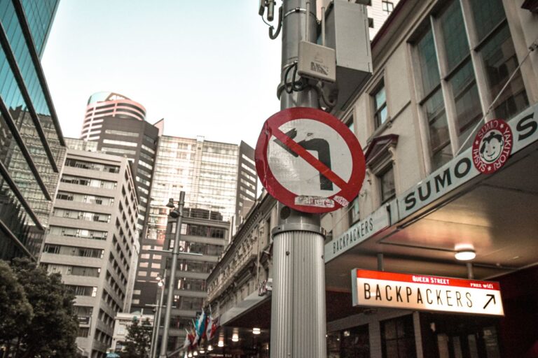 Urban cityscape featuring a no U-turn sign on Queen Street, Selandia Baru, with modern high-rise buildings.